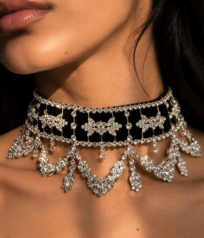 A close-up of a model wearing an ornate silver choker necklace with intricate crystal embellishments, a black velvet band, and cascading crystal fringe.